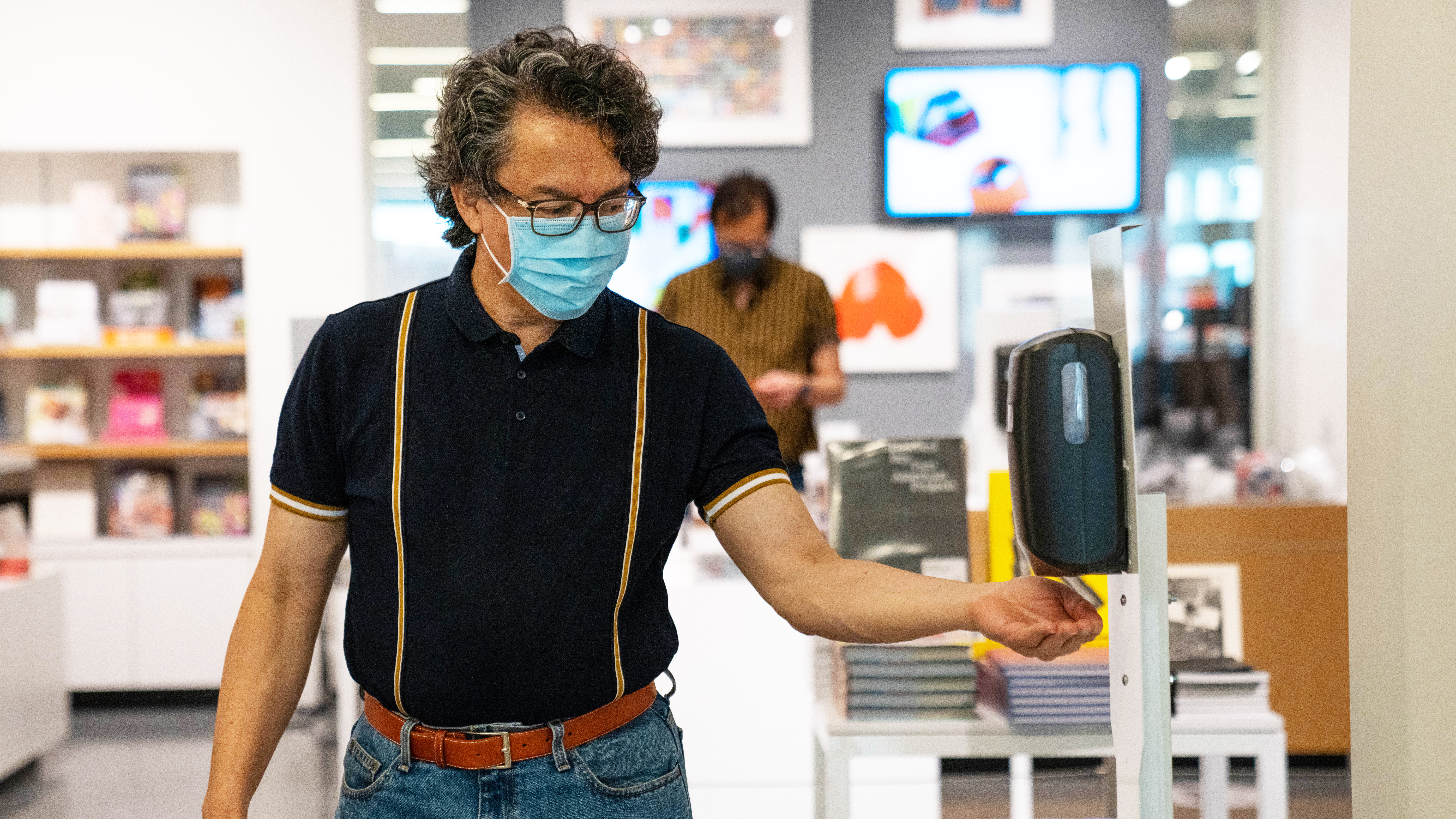 A person wearing a face mask puts hand beneath hand sanitizer dispenser station inside of the SFMOMA museum store.